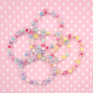 May include: Several colorful beaded bracelets with flower-shaped beads and small round beads in pastel colors. The bracelets are arranged on a pink background with a heart pattern.
