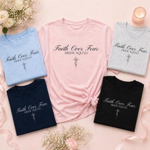 Könnte beinhalten: Fünf T-Shirts in verschiedenen Farben: Hellblau, Rosa, Grau, Marineblau und Schwarz. Jedes Shirt zeigt den Text "Faith Over Fear BRIDE SQUAD" in einer Schreibschrift mit floralem Design. Die Shirts sind mit Blumen und einer Kerze arrangiert.