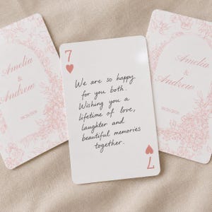 Puede incluir: Tres tarjetas rectangulares con esquinas redondeadas. Dos tarjetas presentan un diseño de arco floral rosa con los nombres Amelia & Andrew y la fecha 08/26/2026. La tarjeta central es un 7 de corazones con un mensaje escrito a mano.