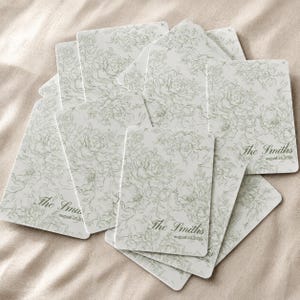 Puede incluir: Un montón de tarjetas rectangulares blancas con un delicado patrón de líneas florales en color verde salvia. Cada tarjeta muestra el texto The Smiths y la fecha August 26, 2026, en una fuente cursiva, dispuestas en un diseño superpuesto.