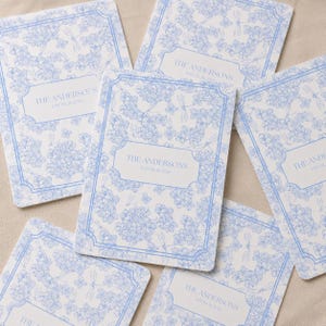 Puede incluir: Una colección de tarjetas rectangulares con un patrón de flores y libélulas en azul claro sobre fondo blanco. Cada tarjeta tiene un marco rectangular blanco central con el texto THE ANDERSONS y EST 06.26.2026 en color azul.