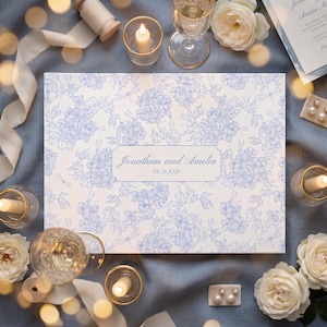 May include: A wedding guest book with a blue floral pattern and the names "Jonathan and Amelia" and the date "06.26.2026" printed on the cover. The book is surrounded by candles, flowers, and ribbons.