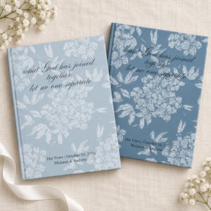 May include: Two hardcover vow books with a floral pattern in shades of blue. The left book is light blue with the text "what God has joined together, let no one separate" and "Her Vows | October 16, 2026 | Melanie & Andrew". The right is dark blue.
