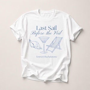 Op de afbeelding: Wit T-shirt met een blauw ontwerp. Het ontwerp bevat een zeeschelp, een martiniglas en een strandstoel. De tekst "Last Sail Before the Veil" en "Emma's Bachelorette" is ook blauw.