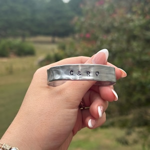 May include: A silver-toned metal cuff bracelet with a hammered, uneven texture. The surface is stamped with the black text C & R followed by a small heart symbol. The bracelet has a wide, flat band design with rounded edges and a metallic finish.