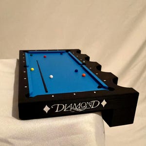 Peut inclure: Un billard miniature noir avec un tapis bleu vif, portant le mot DIAMOND en lettres blanches sur le côté. La table comprend une queue de billard noire et plusieurs petites boules de billard colorées éparpillées sur la surface de jeu.