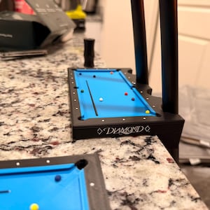 May include: A miniature pool table with a bright blue felt surface and a black frame. The table has a Diamond logo and is set with several billiard balls and a cue stick. The table is on a countertop.