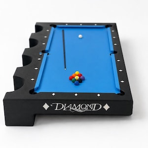 May include: A miniature black pool table with a bright blue felt surface, featuring the word DIAMOND printed on the side. The table includes a black cue stick, a white cue ball, and a small cluster of colorful billiard balls arranged on the surface.