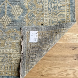 May include: A rectangular rug with a distressed, vintage appearance. The rug features a central design in shades of blue and beige, with intricate geometric patterns. The edges are bordered in a darker blue, and the rug's underside is visible, revealing its construction.