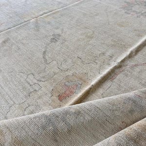 May include: A distressed vintage rug in a neutral colour palette. The rug displays faded floral patterns in muted reds, greens, and greys. The texture of the rug is visible, with a slightly rolled edge, giving it a worn appearance.
