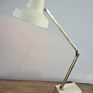 May include: A desk lamp featuring an off-white, textured conical shade and a matching rectangular base. The lamp has a multi-jointed, polished brass-toned metal arm that allows for adjustable positioning, connected by visible silver-toned hardware.