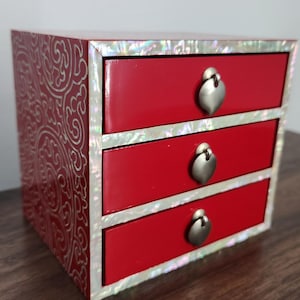 May include: A small, red jewelry box with three drawers. The box has a decorative, swirling pattern on the sides and top. The drawers are trimmed with iridescent mother-of-pearl and have heart-shaped metal pulls.