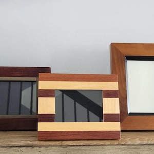 May include: Three wooden picture frames of varying designs. The left frame is dark brown, the center frame features horizontal stripes of light and dark wood, and the right frame is a medium-toned wood with a black inner border and white matting.