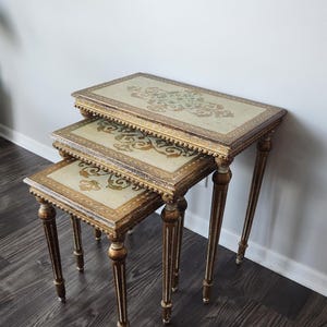 May include: A set of three nesting tables with rectangular tops featuring a cream-colored surface with ornate gold and green floral patterns. The tables have distressed gold-painted wooden frames and slender, tapered legs with carved detailing.