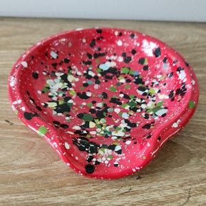 May include: A red ceramic spoon rest with a speckled design in black, white, green, and blue. The spoon rest has a curved shape with a small lip on one side.