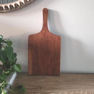 May include: A rectangular wooden cutting board with a long, narrow handle featuring a small circular hole at the top. The board has a smooth, medium-brown finish with visible natural wood grain patterns throughout the surface of the wood.