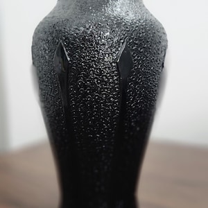 May include: A black glass vase with a textured surface and a glossy finish. The vase has a wide base that tapers upwards, with a narrow neck. The surface has a pattern of small, raised dots. Decorative home decor.