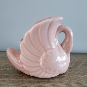 May include: A light pink ceramic swan-shaped planter or vessel featuring molded wing details on the sides, a smooth glossy finish, and a curved neck that forms a handle. The object has a hollow center and a rounded base, showcasing a stylized bird form.