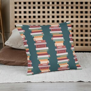 Puede incluir: Un coj&iacute;n decorativo con un fondo verde azulado y un patr&oacute;n repetido de libros apilados. Los libros son de varios colores, incluyendo rojo, naranja y blanco. El coj&iacute;n es rectangular y parece estar hecho de una tela suave.