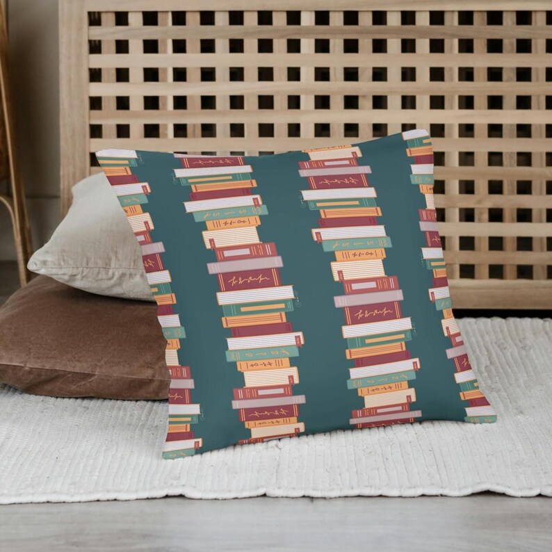 Puede incluir: Un coj&iacute;n decorativo con un fondo verde azulado y un patr&oacute;n repetido de libros apilados. Los libros son de varios colores, incluyendo rojo, naranja y blanco. El coj&iacute;n es rectangular y parece estar hecho de una tela suave.