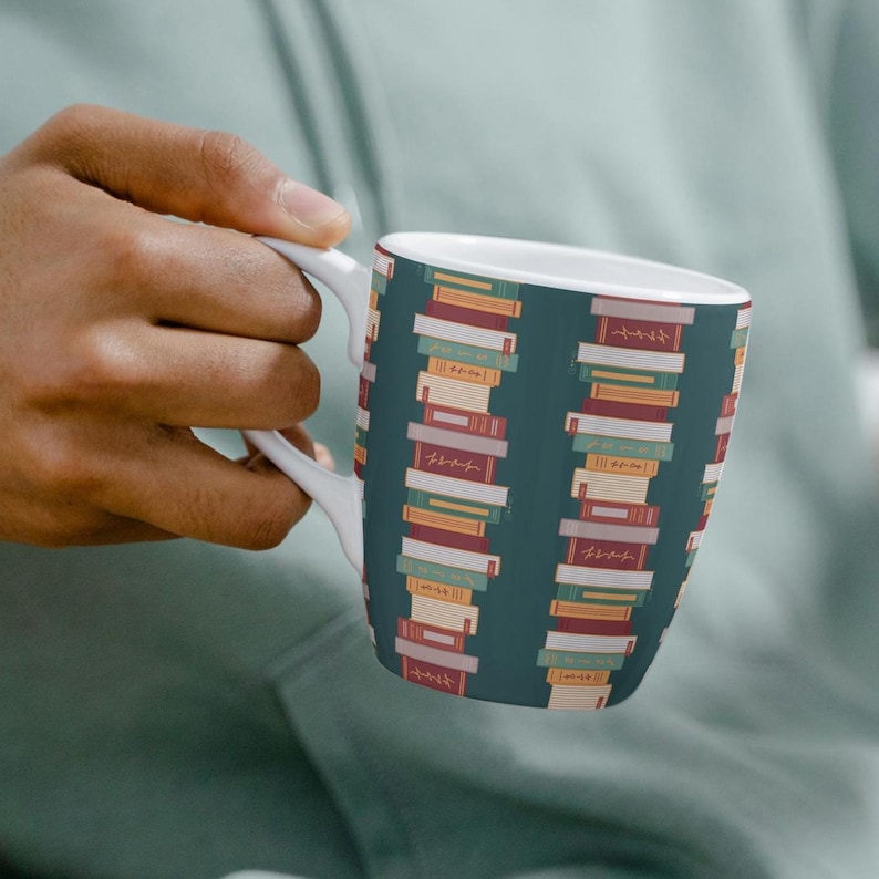 Puede incluir: Taza de cer&aacute;mica blanca con fondo verde azulado y un patr&oacute;n repetido de libros apilados de colores. Los libros tienen varios colores, incluyendo rojo, amarillo y naranja. La taza tiene un asa blanca y la sostiene una persona.