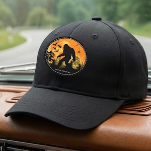 Può includere: Un cappellino da baseball nero con una toppa circolare sul davanti. La toppa mostra uno sfondo al tramonto arancione e giallo con la sagoma nera di una creatura simile a Bigfoot, alberi, pipistrelli e un ragno, con il testo Cryptic Realm Inc.