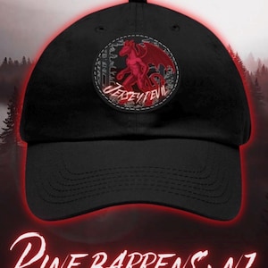 May include: A black baseball cap featuring a circular patch on the front. The patch displays a red winged creature illustration with the text JERSEY DEVIL. Below the cap, the text PINE BARRENS, NJ is written in a stylized white font.