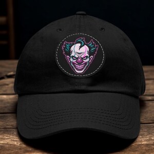 May include: A black baseball cap featuring a circular patch on the front panel. The patch displays a graphic illustration of a clown face with teal hair, a purple nose, and a wide, toothy grin, outlined with white stitching against a black background.