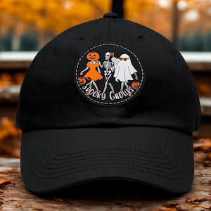 May include: A black baseball cap featuring a circular patch on the front. The patch displays a pumpkin-headed figure in an orange dress, a skeleton, and a white ghost wearing sunglasses. The text "SPOOKY GHOULS" is printed below the figures.