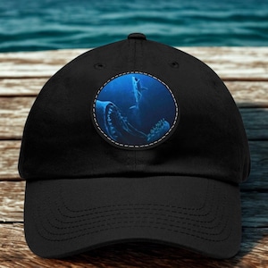 May include: A black baseball cap featuring a circular patch on the front. The patch displays a deep blue underwater scene with a large shark silhouette and a smaller shark swimming above it, surrounded by a white stitched border on the cap fabric.