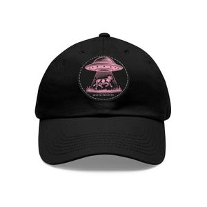 Peut inclure: Une casquette de baseball noire avec un écusson circulaire sur le devant. L'écusson présente un motif rose d'un OVNI enlevant une vache dans un champ d'herbe, avec le texte "CRYPTID REALM INC." imprimé en petites lettres blanches en bas.