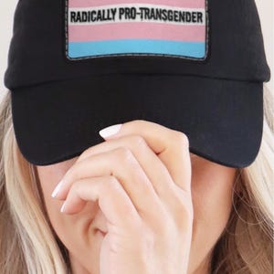 Puede incluir: Una gorra de béisbol negra con un parche rectangular en la parte delantera. El parche muestra los colores de la bandera del orgullo transgénero en azul claro, rosa y blanco, con el texto RADICALLY PRO-TRANSGENDER en letras mayúsculas negras.