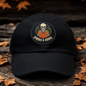 Op de afbeelding: Een zwarte honkbalpet met een ronde patch op de voorkant. De patch toont een skelet dat een open oranje boek vasthoudt, omringd door groene bladeren en kleine sterretjes. De tekst SPOOKS & BOOKS staat in een crèmekleur onder het boek.
