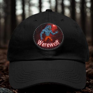 Puede incluir: Una gorra de béisbol negra con un parche circular en la parte delantera. El parche muestra un personaje de hombre lobo azul con pantalones naranjas sobre un fondo de bosque rojo oscuro, con la palabra Werewolf escrita en blanco abajo.