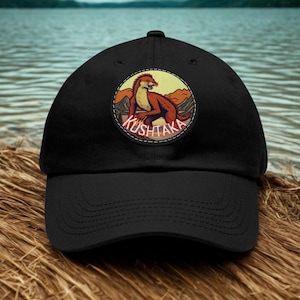 May include: A black baseball cap featuring a circular patch on the front. The patch displays a brown, otter-like creature standing in a mountainous landscape with a yellow sky. The word KUSHTAKA is printed in white letters at the bottom of the patch.