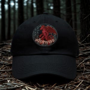 May include: A black baseball cap featuring a circular patch on the front. The patch displays a red winged creature illustration against a dark forest background with the text JERSEY DEVIL written in a stylized font across the lower portion.