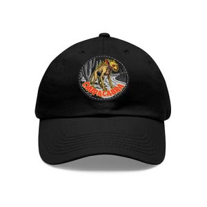 May include: A black baseball cap featuring a circular patch on the front. The patch displays a yellow, spiky-backed creature with red eyes standing in a desert landscape with cacti. The word CHUPACABRA is printed in bold red letters below the creature.