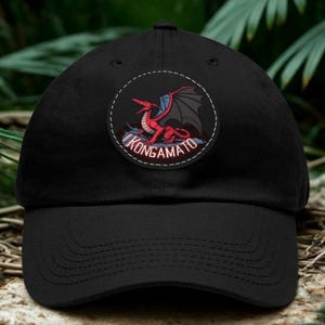 Peut inclure: Une casquette de baseball noire avec un écusson circulaire sur le devant. L'écusson représente une créature ailée rouge aux ailes gris foncé avec le texte KONGAMATO en lettres capitales blanches, le tout entouré d'une bordure cousue.
