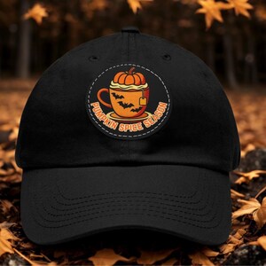 Op de afbeelding: Een zwarte baseballpet met een ronde patch op de voorkant. De patch toont een oranje mok gevuld met een pompoen en een toefje room, versierd met zwarte vleermuissilhouetten. De tekst PUMPKIN SPICE SEASON staat in een cirkel rond de mok.