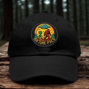 May include: A black baseball cap featuring a circular patch on the front. The patch displays a graphic of an alien and a Bigfoot walking together under a flying saucer against a yellow sun and green trees, with the text NATURE PALS below.