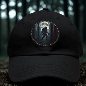 Op de afbeelding: Een zwarte honkbalpet met een ronde patch op het voorpaneel. De patch toont het donkere silhouet van een wezen dat op Bigfoot lijkt, lopend door een bos in de nacht, tegen de achtergrond van een grote, heldere volle maan.