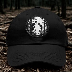 Può includere: Un cappellino da baseball nero con una toppa circolare sul pannello frontale. La toppa mostra un'illustrazione grafica in bianco e nero di due figure aliene snelle in piedi all'interno di una fitta foresta, circondate da alberi.