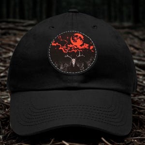 May include: A black baseball cap featuring a circular patch on the front. The patch displays a graphic illustration of a deer skull with antlers set against a dark forest background, topped with red clouds and a large, glowing red moon in the sky.