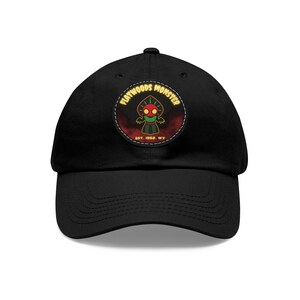 May include: A black baseball cap featuring a circular patch on the front. The patch displays a cartoon character with a red head and yellow eyes, surrounded by the text "FLATWOODS MONSTER" and "EST. 1952, WV" against a dark red background.