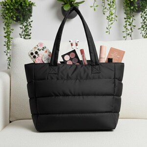 May include: A black quilted tote bag filled with beauty products, a floral phone case, and wireless earbuds. The bag has two handles and is sitting on a white couch. The background features hanging plants.