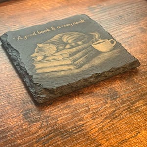 May include: A square slate coaster with rough, uneven edges featuring an engraved illustration of a cat sleeping on a stack of books next to a steaming mug. The text "A good book & a cozy nook" is engraved in a script font above the illustration.