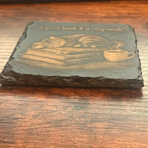 May include: A square slate coaster with a rough, natural edge featuring an engraved illustration of a cat sleeping on a stack of books next to a steaming cup of coffee. The text "A good book & a cozy nook" is engraved at the top of the coaster.