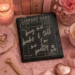 May include: A rectangular black slate plaque featuring a white printed library card design with the text "LIBRARY CARD", "CARD NUMBER 026483", and the handwritten-style phrase "buy me books & tell me I'm pretty" with three small heart icons.