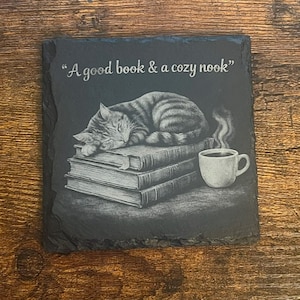 May include: A square black slate coaster with rough, uneven edges. The surface features an engraved illustration of a tabby cat sleeping on a stack of three books next to a steaming mug. The text "A good book & a cozy nook" is engraved above.