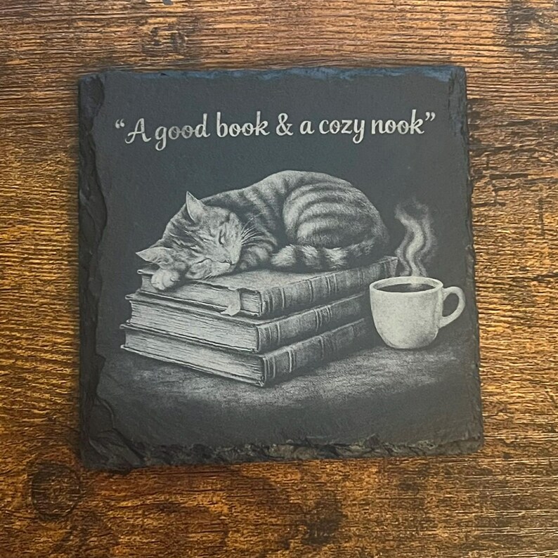 May include: A square black slate coaster with rough, uneven edges. The surface features an engraved illustration of a tabby cat sleeping on a stack of three books next to a steaming mug. The text "A good book & a cozy nook" is engraved above.
